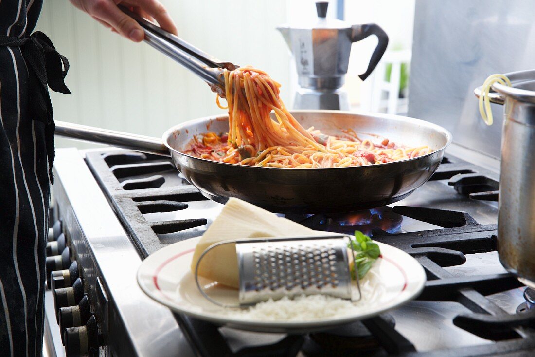 A person preparing linguine with tomato sauce in a pan on a stove