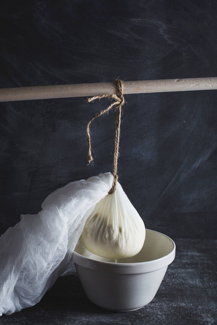 Ricotta in a cheesecloth hanging from a wooden stick and draining into a bowl