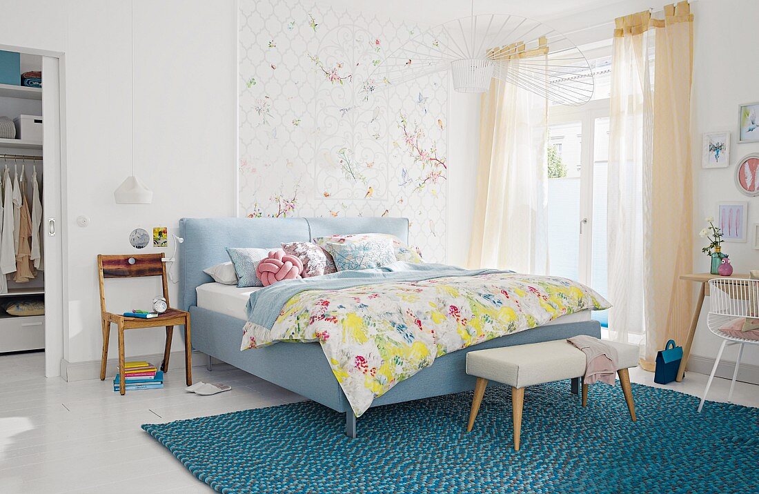 A padded double bed and bench on a turquoise rug in a light, bright bedroom