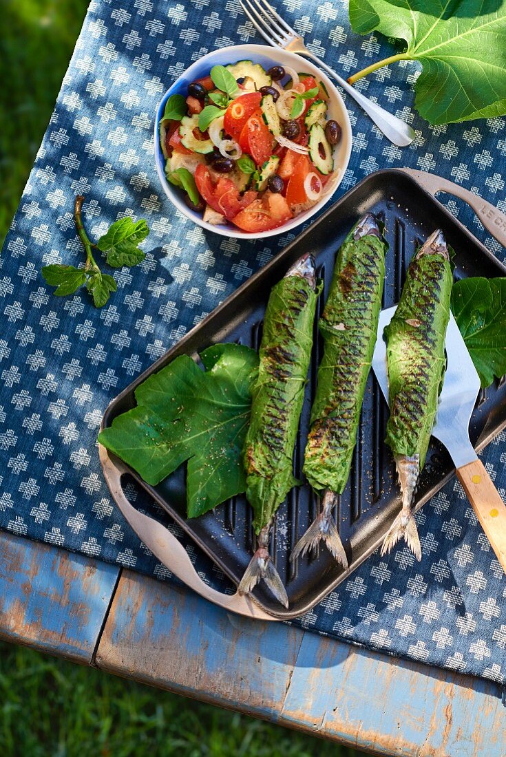 Grilled mackerel wrapped in fig leaves with a bread salad on a table outdoors