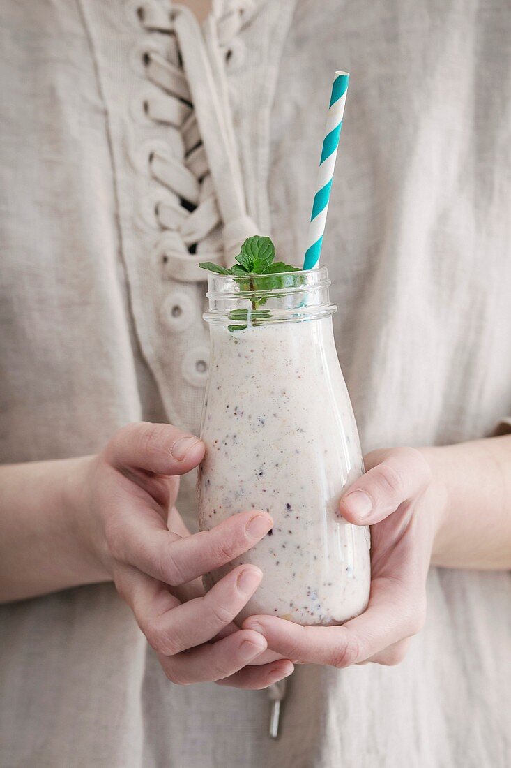 Bottle of smoothie with chia seeds, blueberries, mint leaves and retro striped cocktail tube in female hands