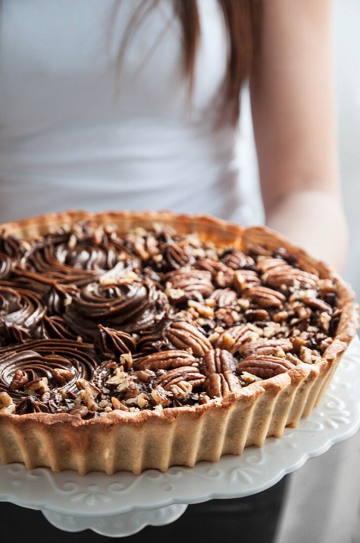 A woman holding a chocolate and pecan tart on a cake stand