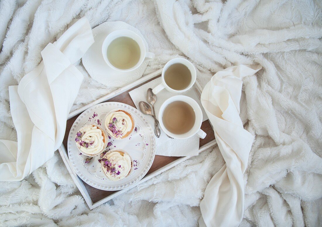 Vanille-Cupcakes mit Tee auf Tablett und weisser Kuscheldecke (Aufsicht)