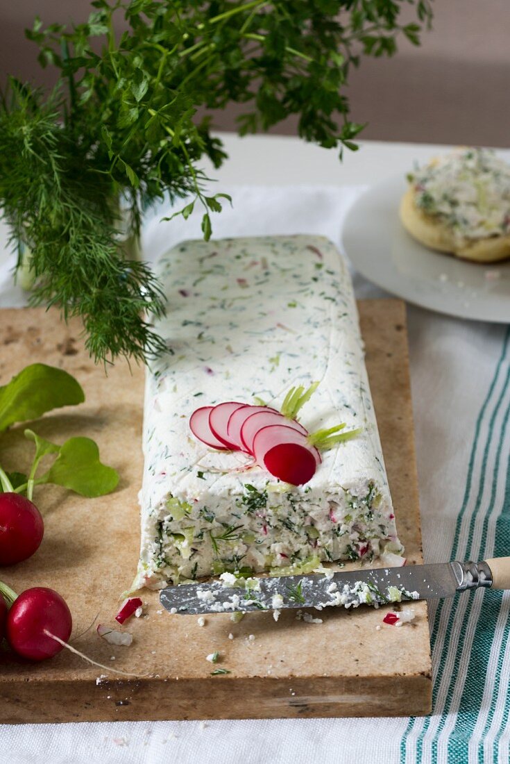A big piece of quark cheese and herbs terrine on a marble board. Radishes, dill, parsley and a roll on a white plate in the background
