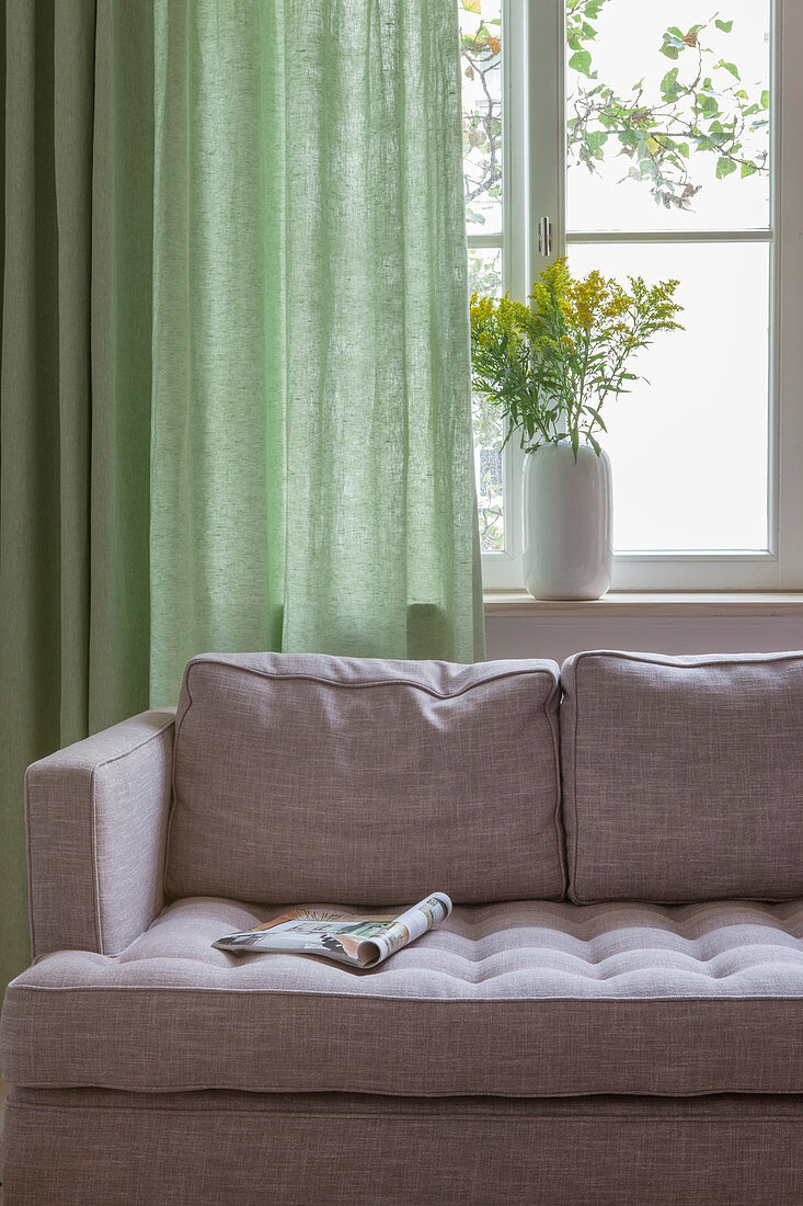 Magazine on grey sofa in front of window with green curtain