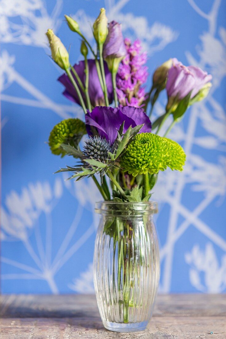 Arrangement of purple flowers in glass vase in front of wall with floral pattern