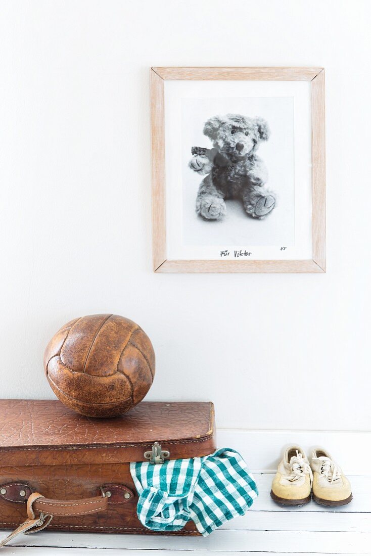 Picture on wall above teddy bear, old ball, leather suitcase and children's shoes