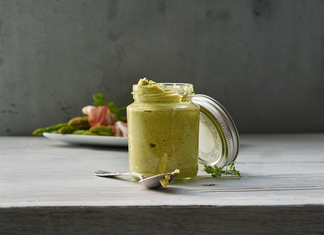 A glass jar of asparagus pesto, with green asparagus and ham in the background