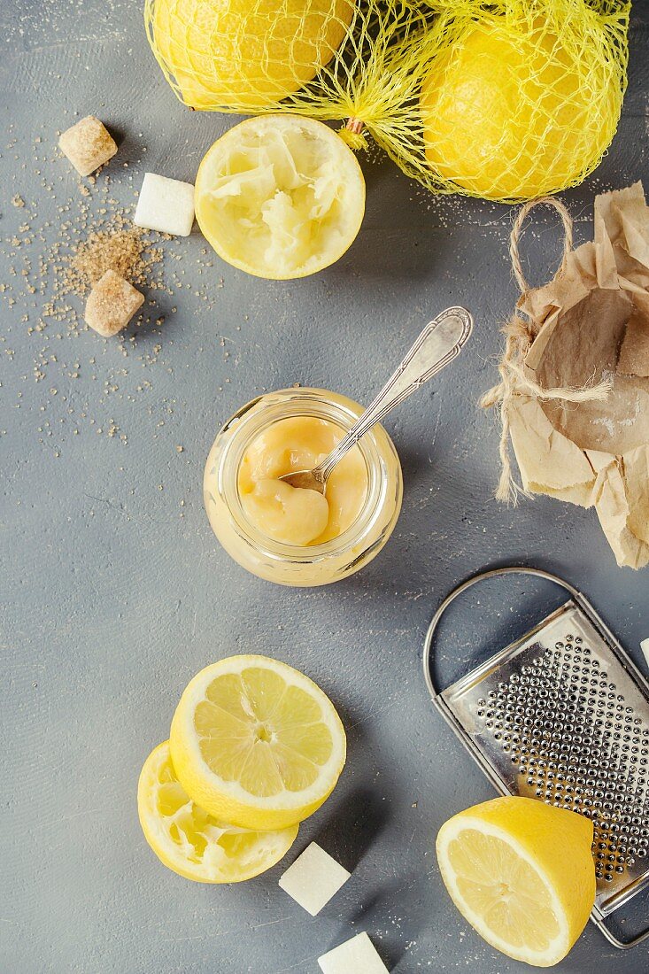 Jar of homemade lemon curd with spoon, whole and sliced lemons, sugar, grater and zest over gray blue stone background