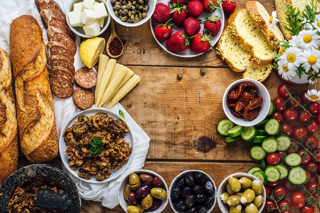 Olive tapenade, sausages, baby corns, french baguette, cheese, capers, strawberries, corn bread slices, sun-dried tomatoes, cherry tomatoes and cucumber slices on a wooden board