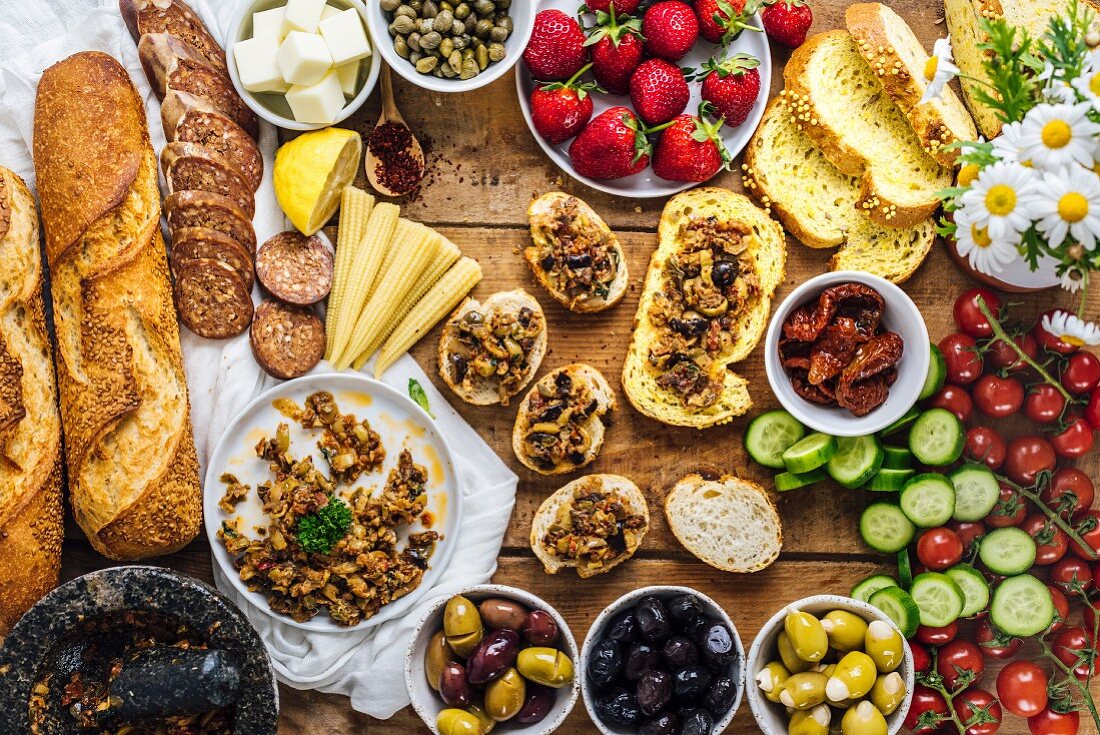 Olive tapenade spread on bread slices served on a wooden board, French baguette, sausages, baby corns, cheese, strawberries