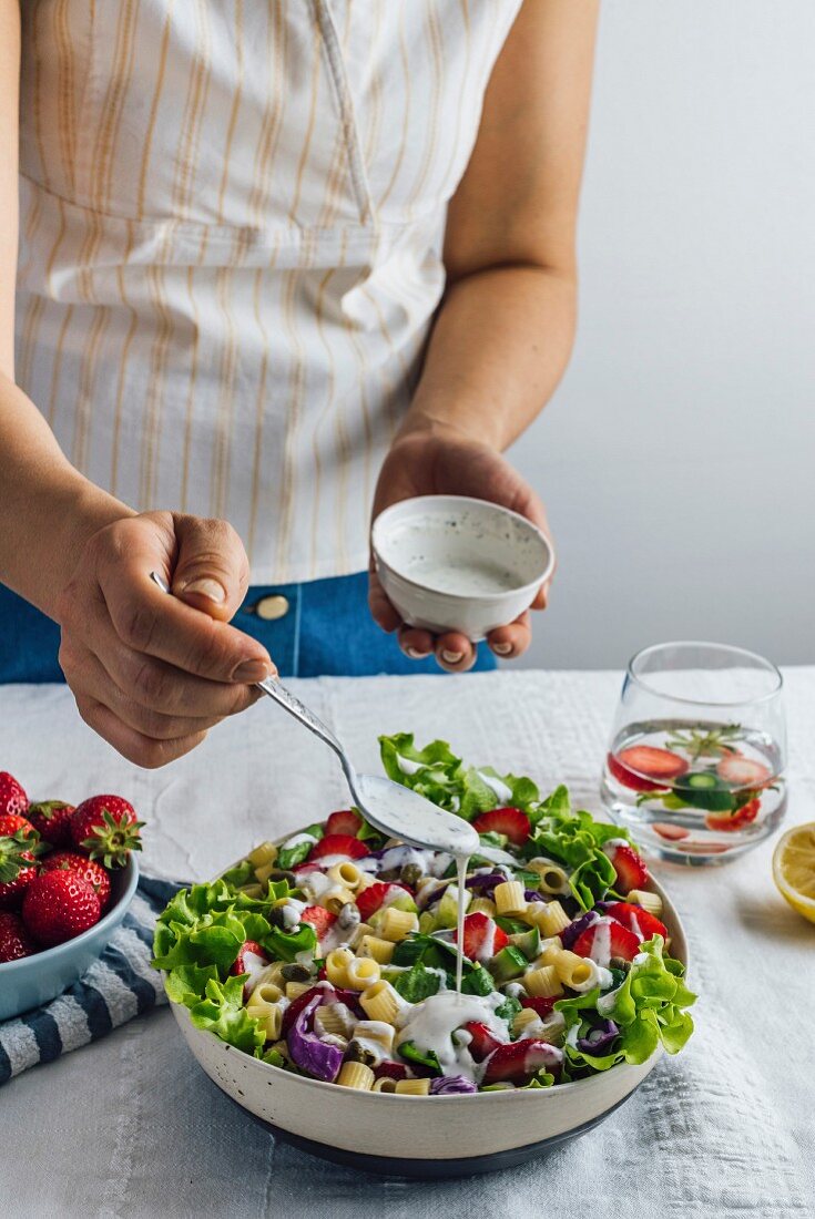 A woman drizzling yogurt dressing over a fresh strawberry pasta salad