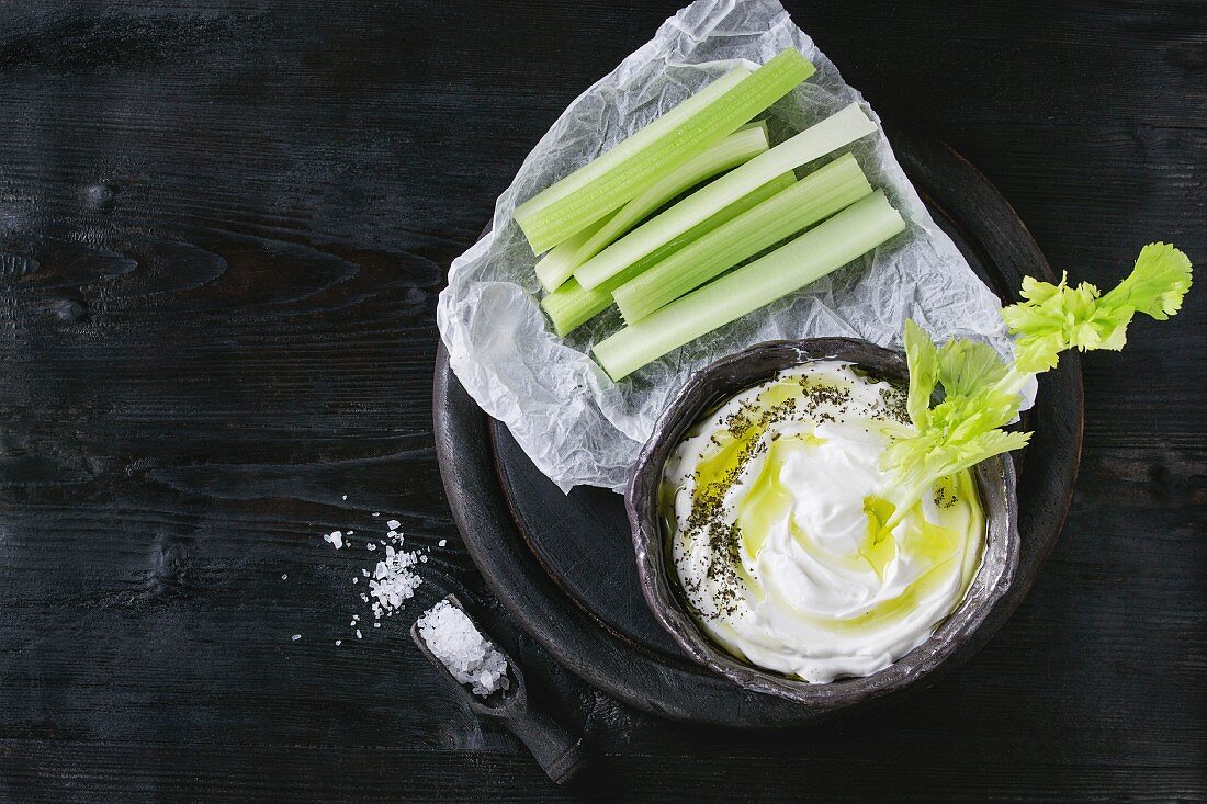 Fresh celery on paper with yogurt and olive oil dip in ceramic bowl, served with sea salt on wood round chopping board