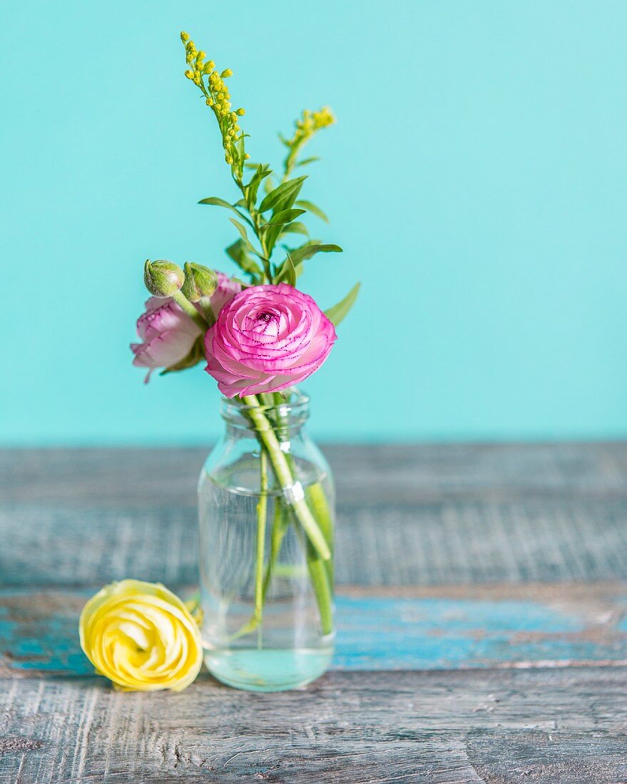 Tiny glass vase of ranunculus on rustic wooden surface against turquoise background