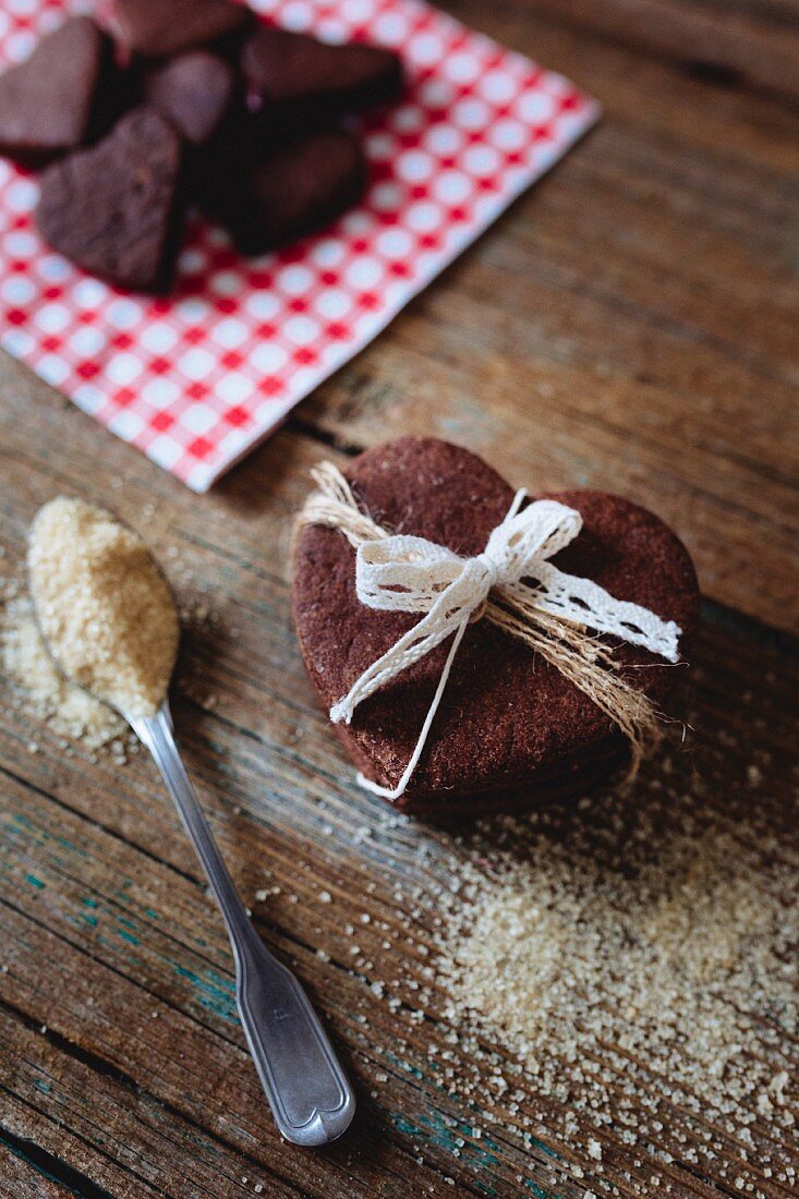 Herzförmige Schokoladenkekse, gestapelt und zusammengebunden, Teelöffel mit braunem Zucker auf Holzuntergrund