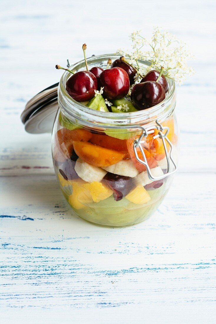 Fruit salad in jar with grapes, mango, banana, cherries, apricot, kiwi, garnished with elder flowers