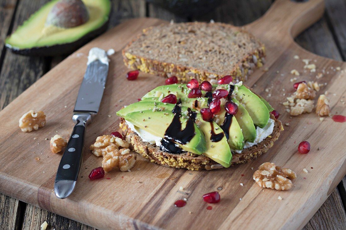 Protein bread slice with cream cheese, sliced avocado, walnuts and pomegranate seed on wooden board