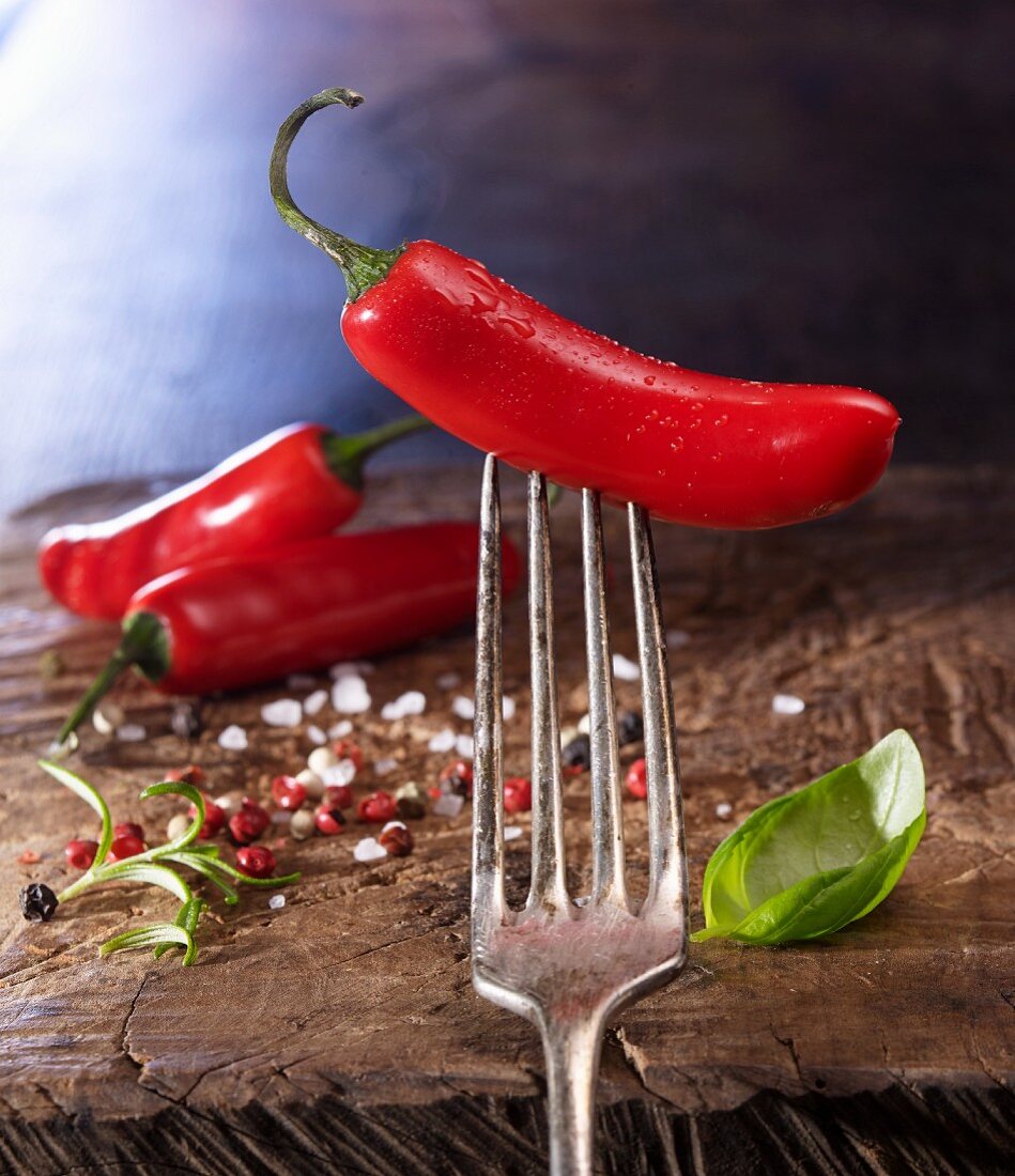 A red chilli pepper on a fork and coloured peppercorns on a wooden board