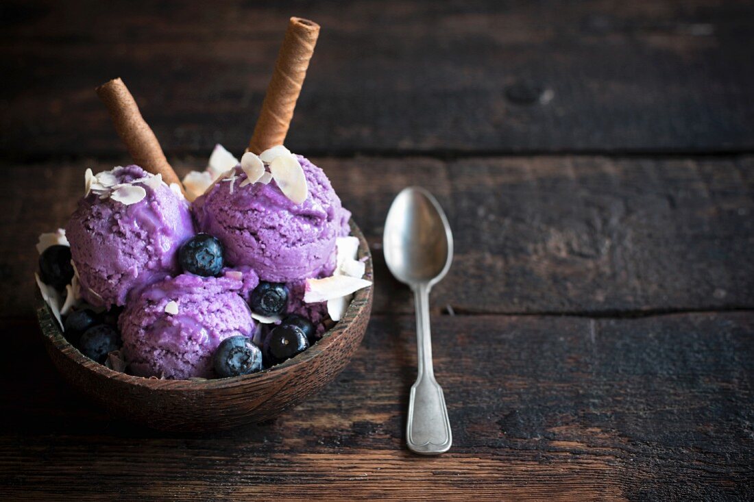 Scoops of blueberry ice cream topped with flaked almonds and chocolate wafer rolls in a wooden bowl