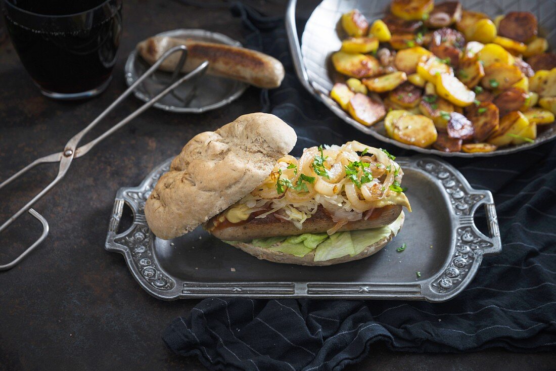 A bread roll with vegan bratwurst, white cabbage and roasted onions, with a plate of fried potatoes