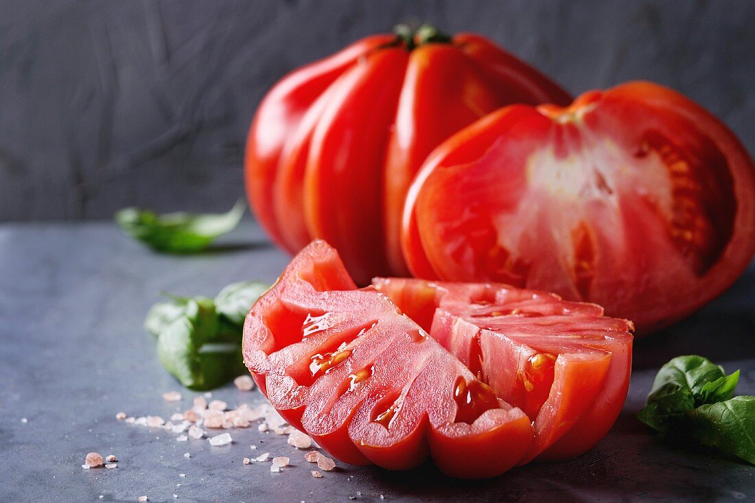 Whole and sliced organic tomatoes Coeur De Boeuf with pink salt and basil on blue gray metal texture background