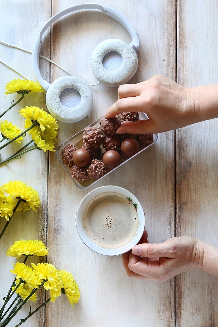 Hands holding a cup of coffee and reaching for a chocolate on a table with flowers and headphones