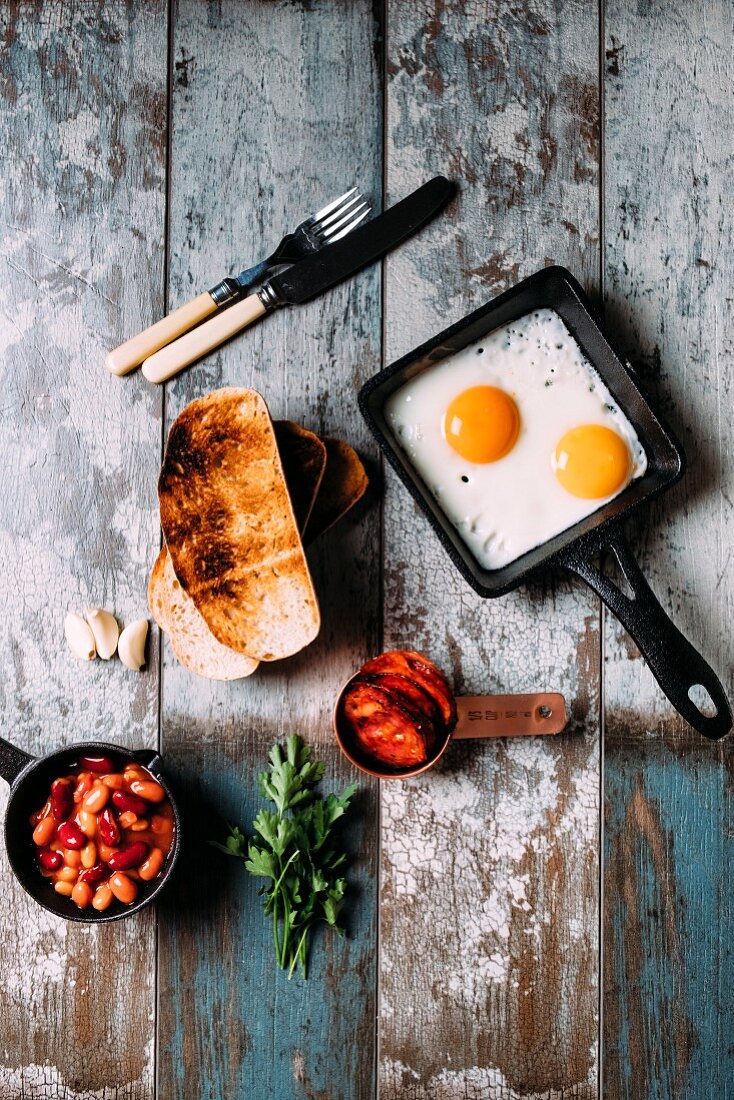 Frühstück mit gemischten Bohnen, getoastetem Sauerteigbrot, Spiegeleiern, Chorizo, Knoblauch und Petersilie