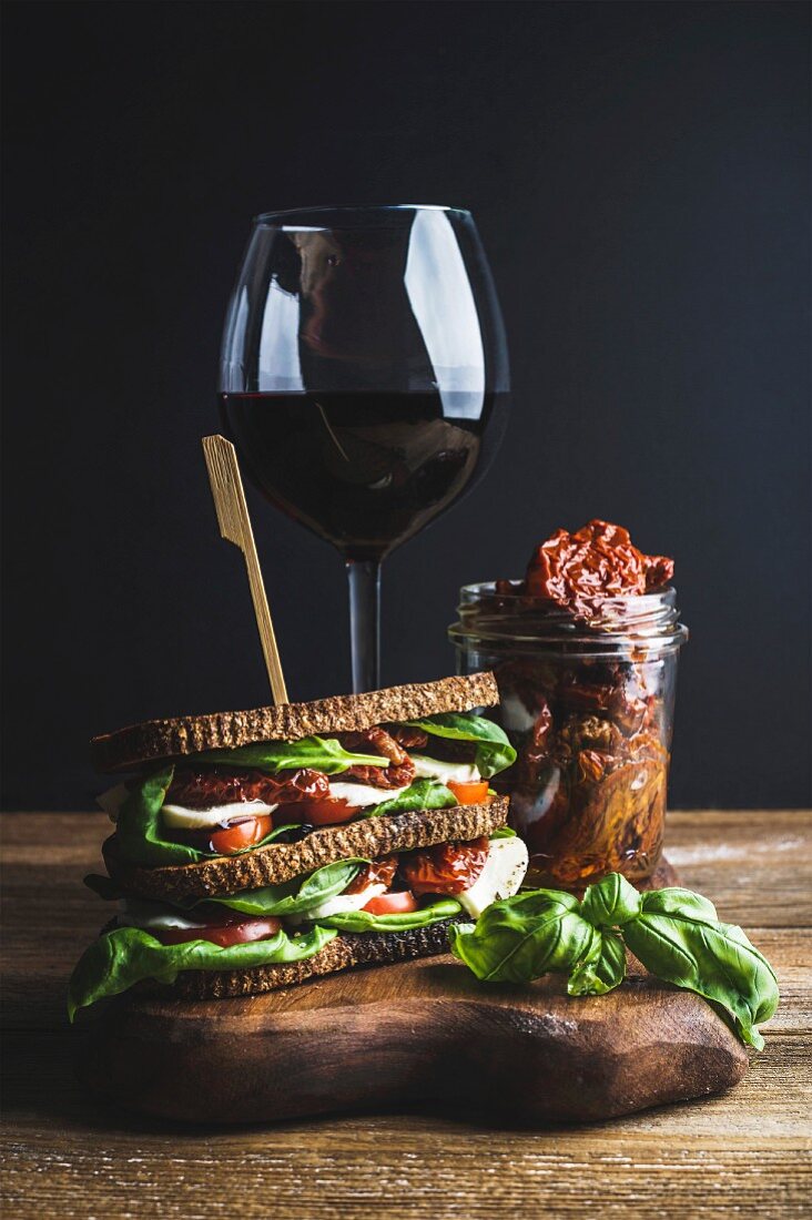 Homemade caprese sandwich, glass of red wine and dried tomatoes in jar on wooden board, dark background