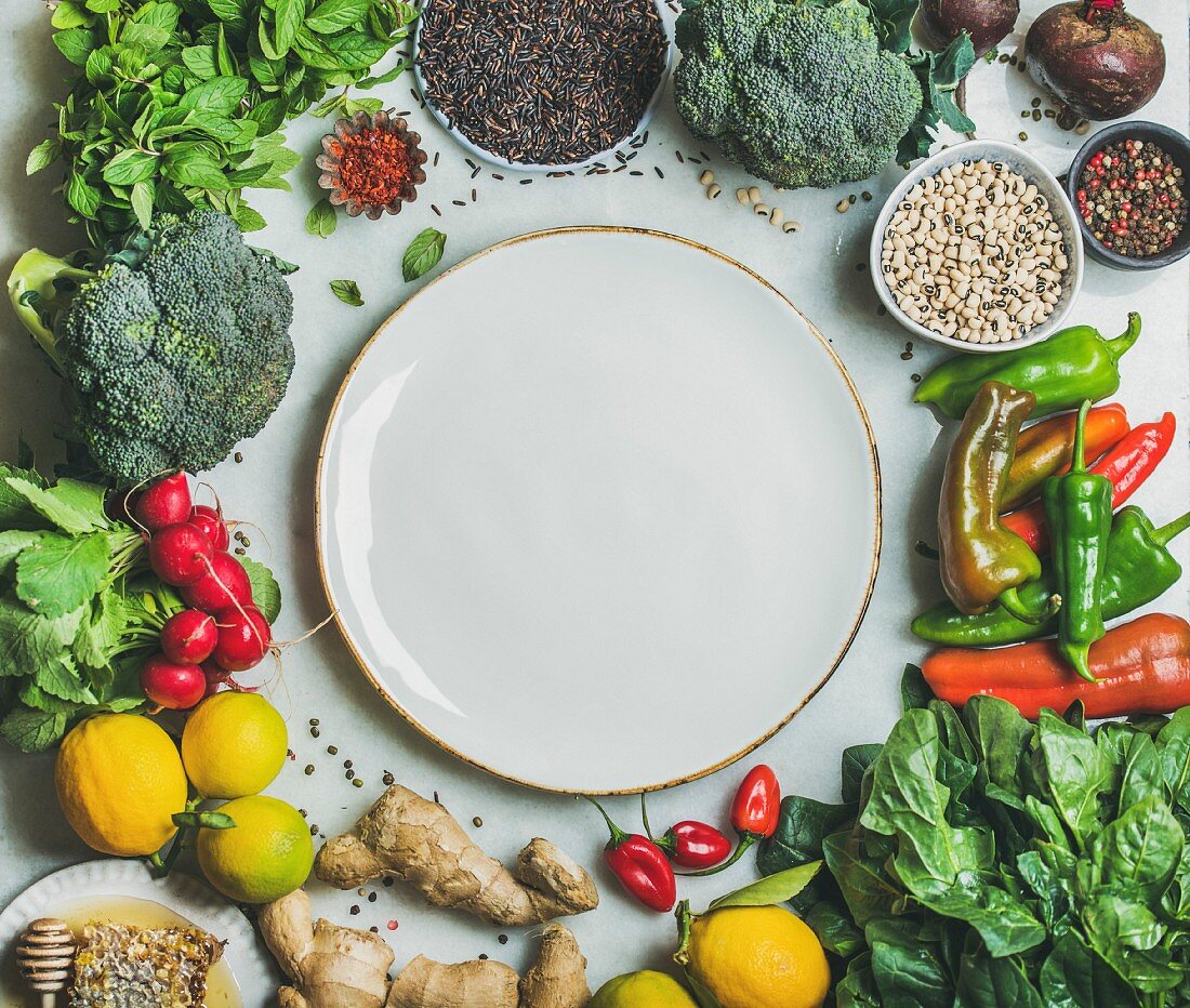 Clean eating healthy cooking ingredients: Vegetables, beans, grains, greens, fruit, spices over grey marble background, white plate with copy space in center