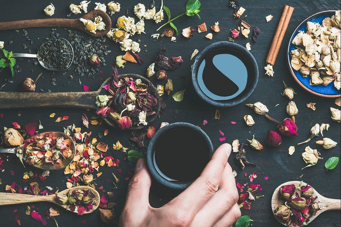 Chinese black tea in black stoneware cups, man's hand holding one cup and wooden spoons with dry herbs
