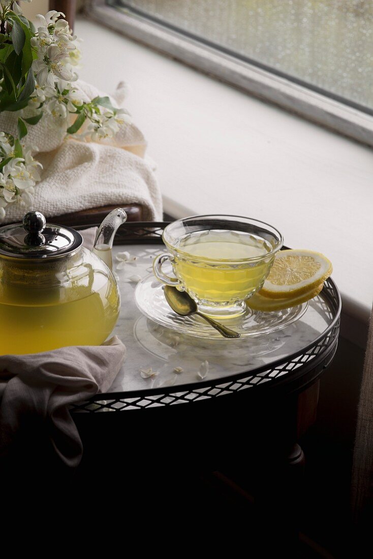 Zitronen-Ingwertee mit Honig in Glaskanne und Tasse auf Tablett am Fenster