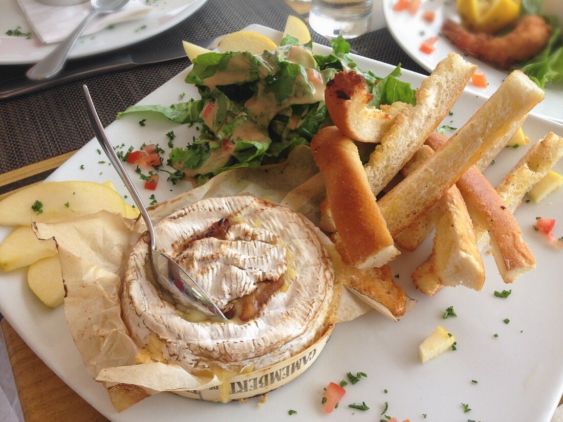 A small wheel of roast Camembert Cheese with toasted bread, apple slices and salad