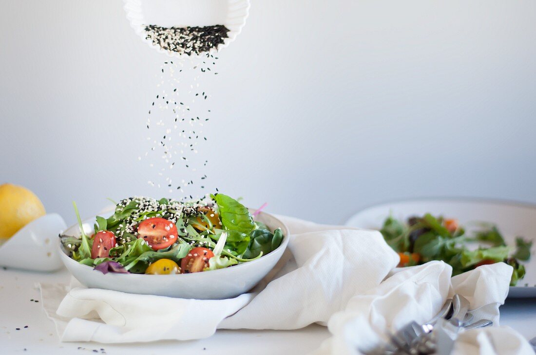 Sesame seeds falling on to a spinach salad with rocket, tomatoes, olive oil, and lemon juice