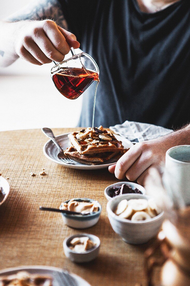 A person pouring syrup on wholemeal waffles with bananas and peanut butter