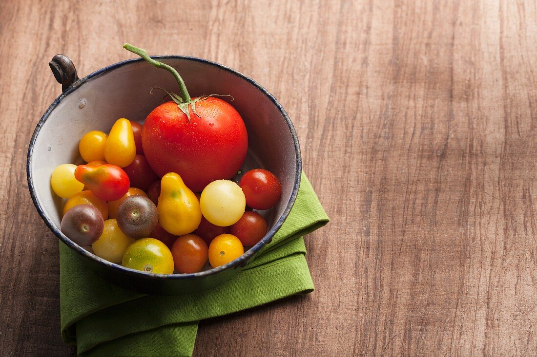 A variety of tomatoes in an antique enamel bowl on wood background with a green napkin