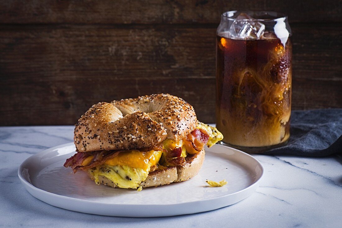 Bagel-Sandwich mit Speck, Rührei und Käse serviert mit einem Glas Eistee