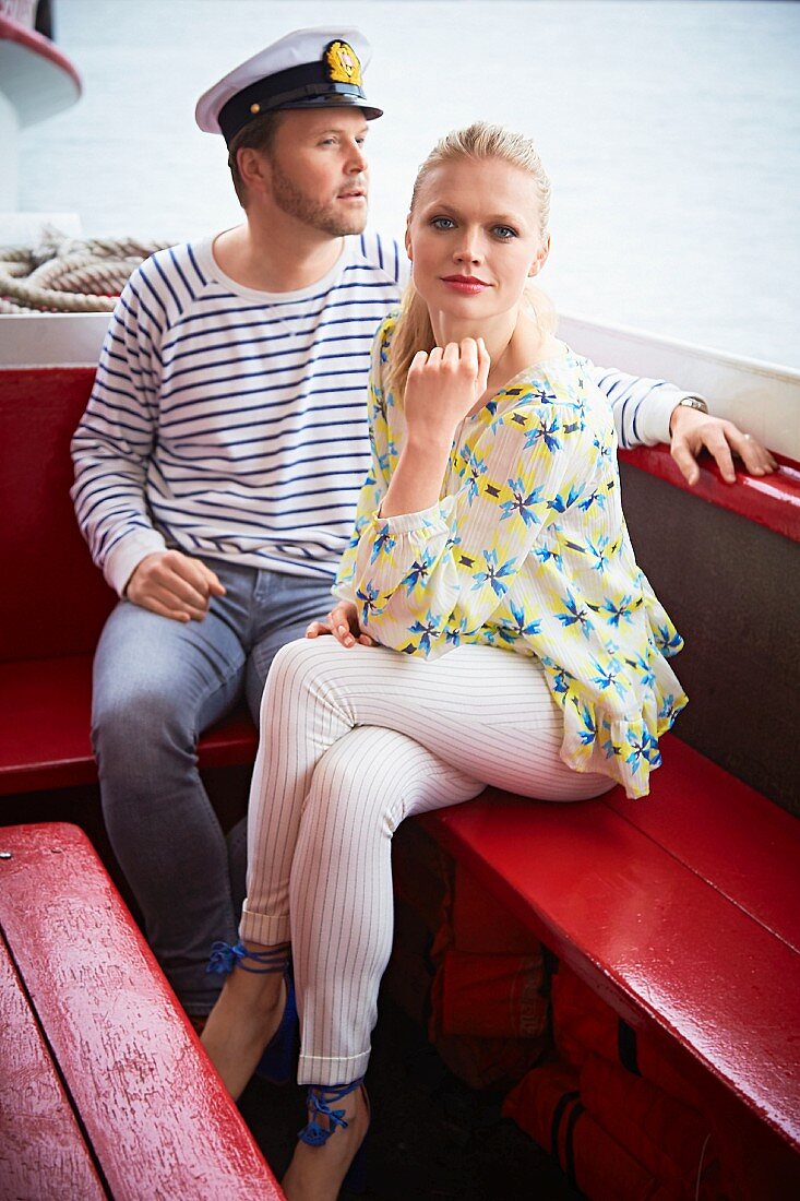 A blonde woman wearing a floral print blouse and white pinstripe trousers with a man in a sailor look on a boat