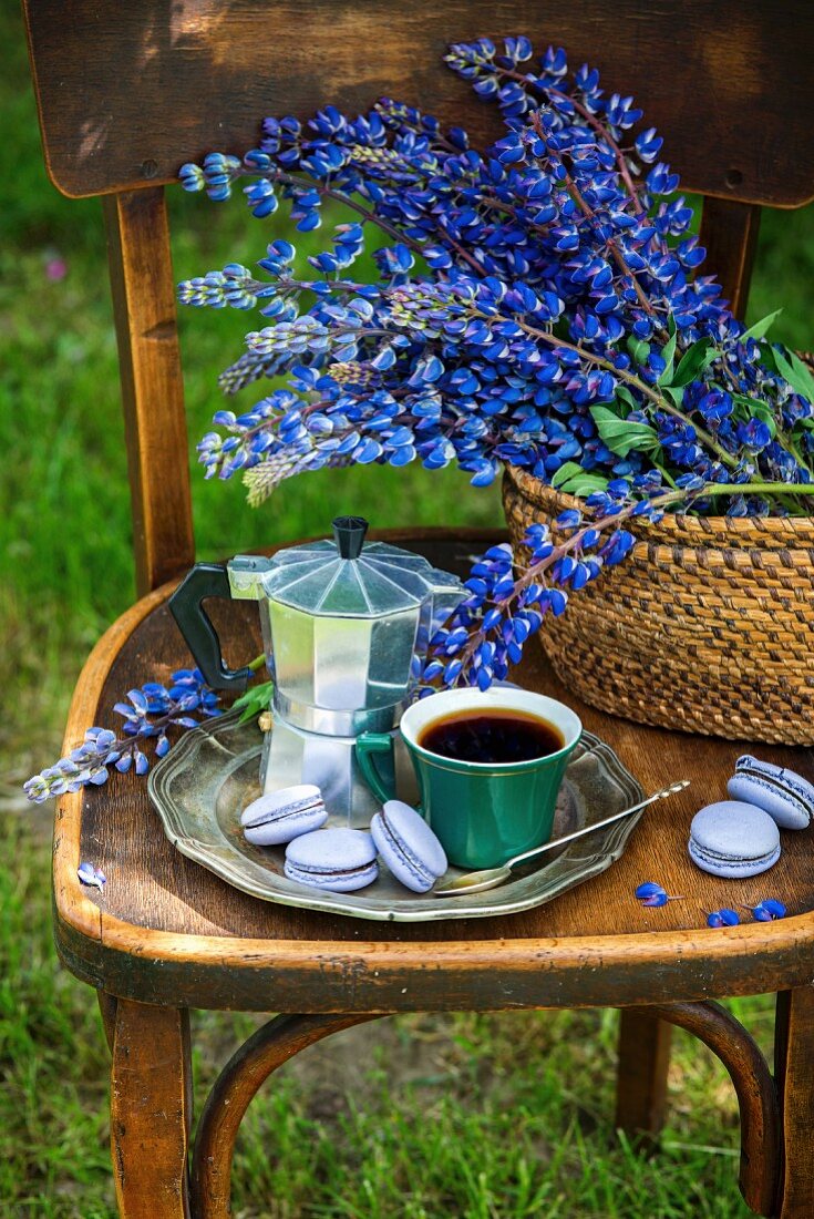 Tasse Kaffee, Macarons und Espressokanne, im Hintergrund Lupinen auf Stuhl im Garten