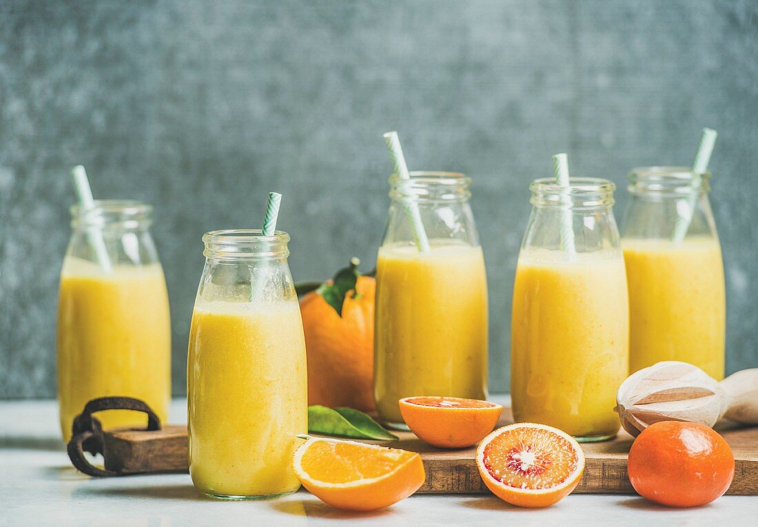 Healthy yellow smoothie with citrus fruit and ginger in bottles on rustic wooden board over light marble table
