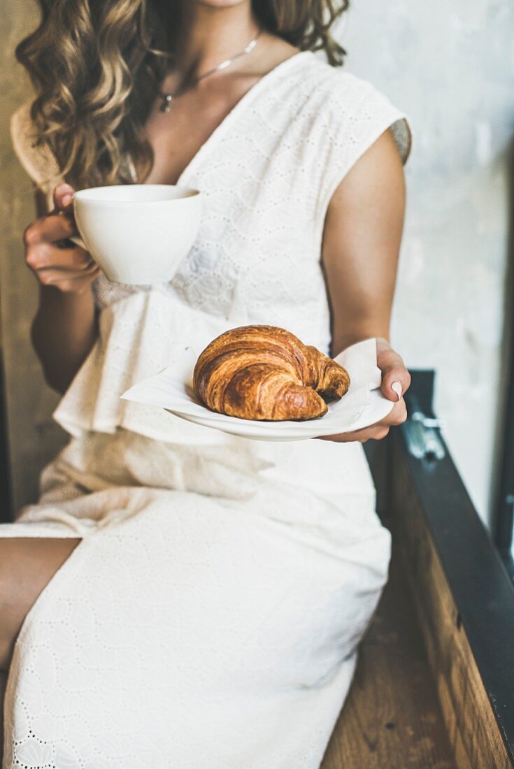 Young blond woman in white dress holding fresh croissant in plate and cup of cappuccino in cafe