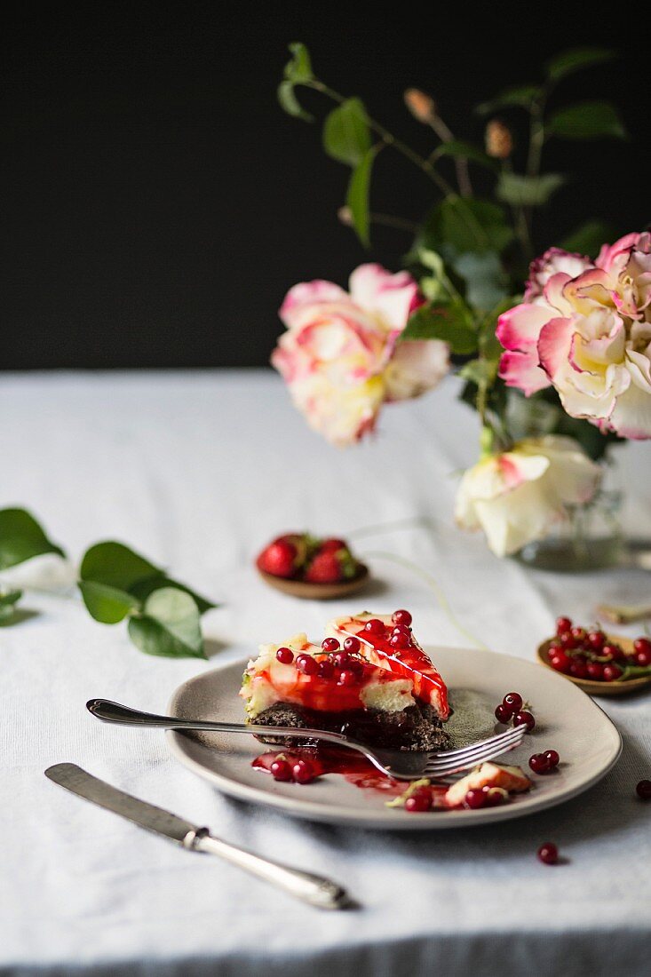 Ein Stück Käsekuchen mit roten Johannisbeeren auf Tisch, daneben Rosenblumenstrauss