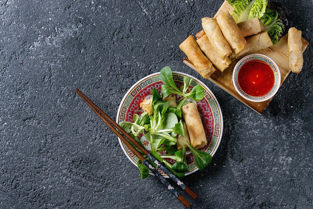 Fried spring rolls with red pepper sauces, served in traditional china plate with fresh green salad and wooden chopsticks over black texture background