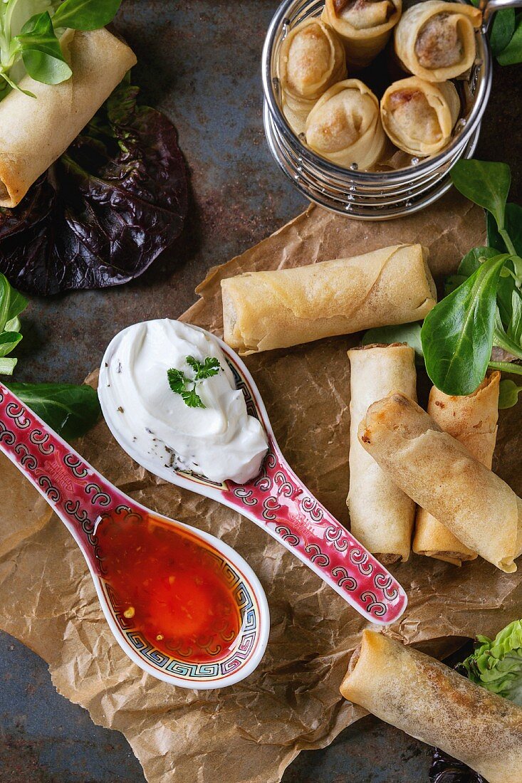 Fried spring rolls with red and white sauces in china spoons, served on crumpled paper and in fry basket with green salad and wooden chopsticks