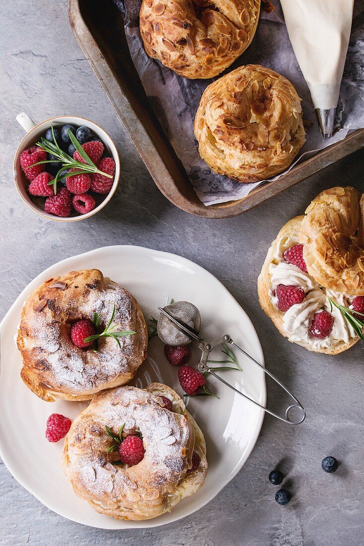 Filling and empty homemade choux pastry cake Paris Brest with raspberries, almond, sugar powder, rosemary on plate and oven tray with berries