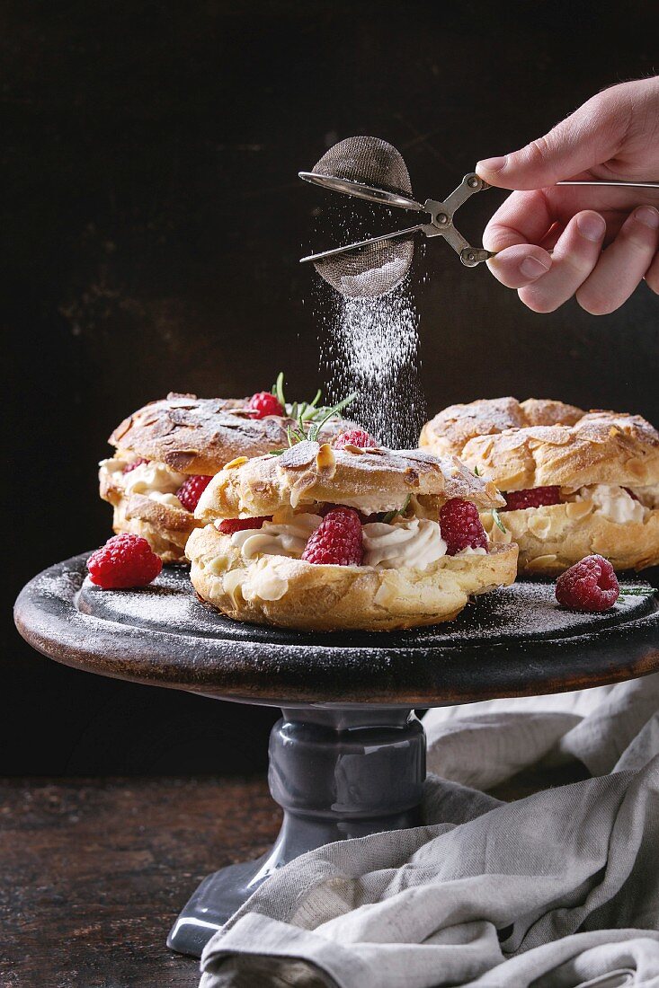 Homemade choux pastry cake Paris Brest with raspberries, almond and rosemary, served on black wooden serving board on cake stand