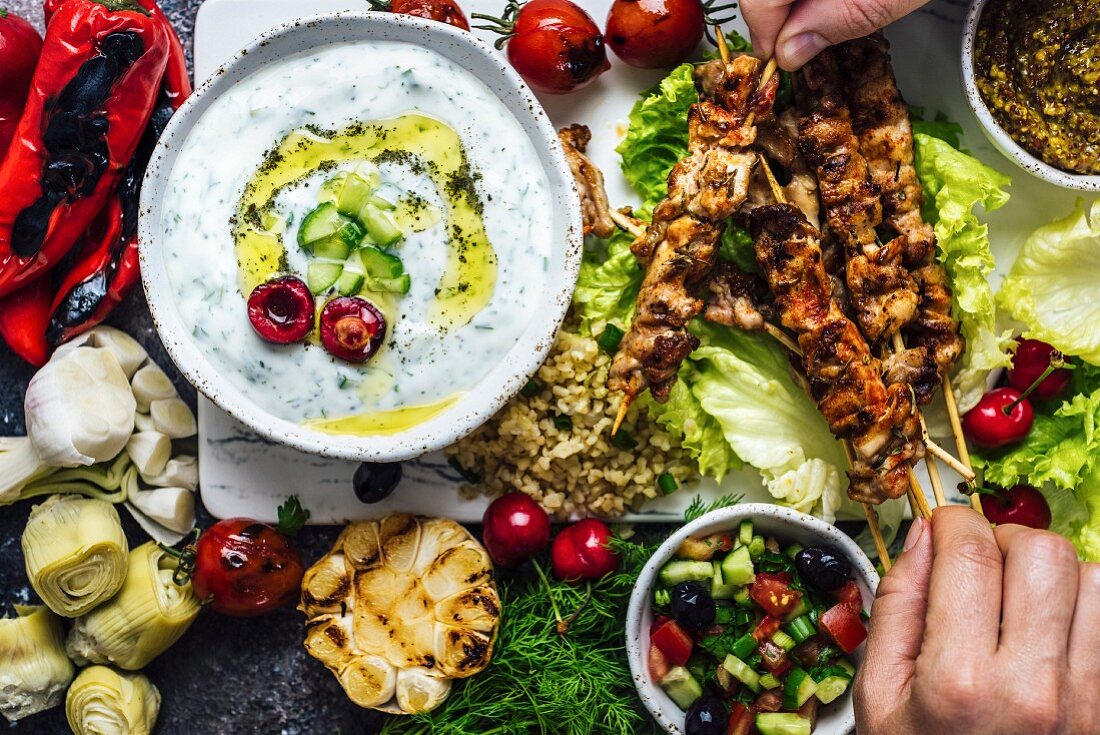 Man and woman getting chicken skewers from a board with lots of snacks and a bowl of yogurt and cucumber dip