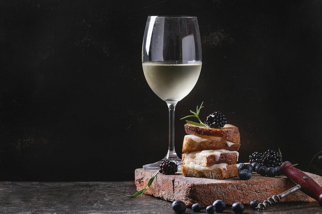 Grilled sandwich with melted goat cheese, blackberry, blueberry, rosemary and honey, served on terracotta board with glass of cold white wine