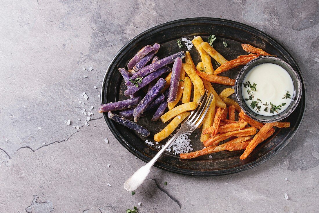 Variety of french fries traditional potatoes, purple potato, carrot served with white cheese sauce, salt, thyme on vintage tray