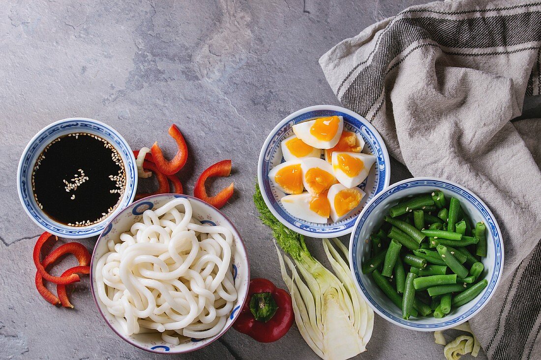 Ingredients for cooking stir fry udon noodles, green beans, sliced paprika, boiled eggs, soy sauce with sesame seeds in traditional bowls with wooden chopsticks