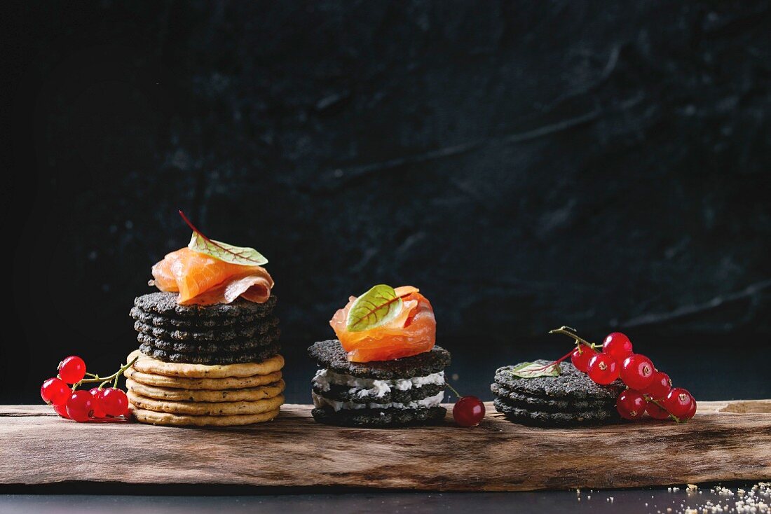 Stack of black charcoal and traditional crackers with smoked salmon, cream cheese, green salad and red currant berries on ood serving board